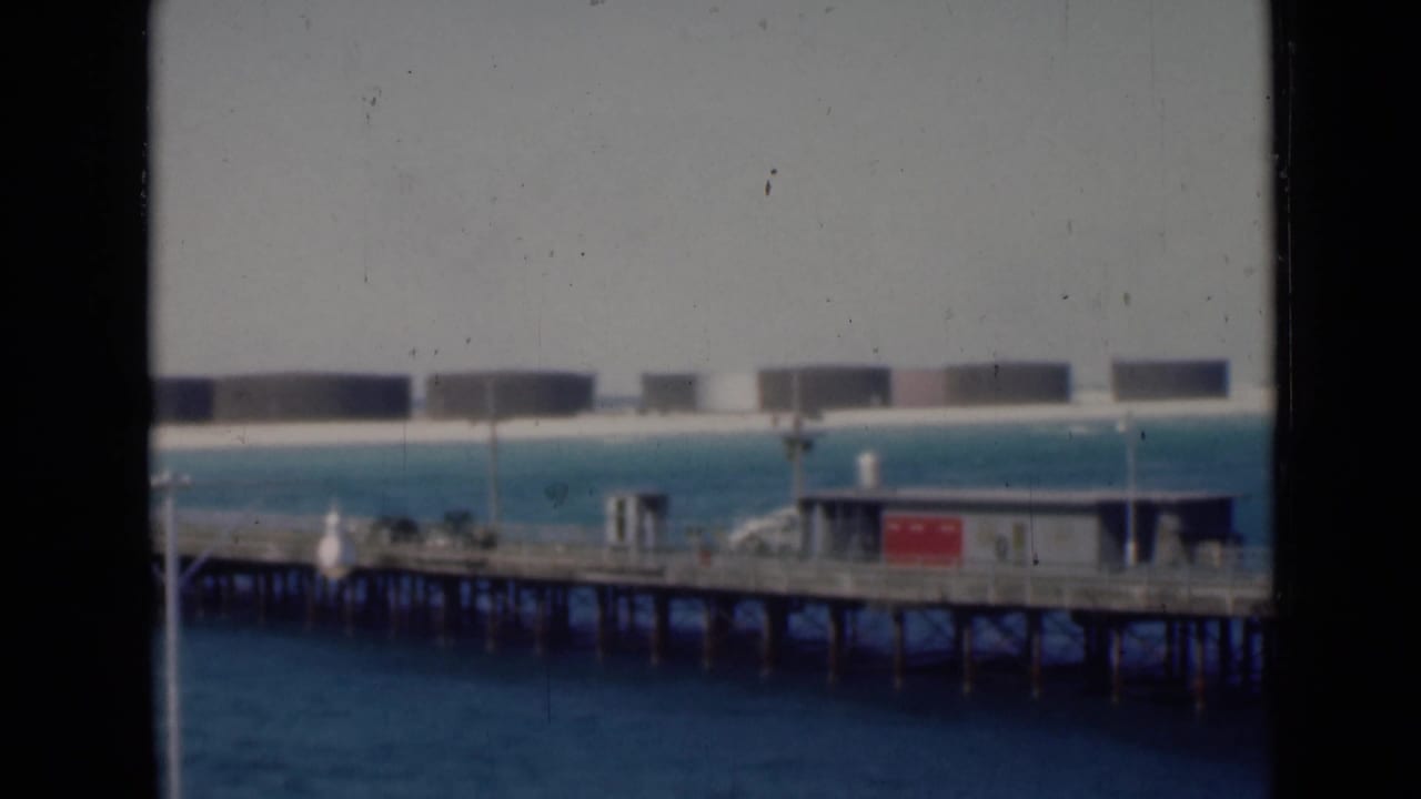 Archival 8mm view of a pier with small structures and a red sign-like panel, with large storage tanks lining the distant shore.