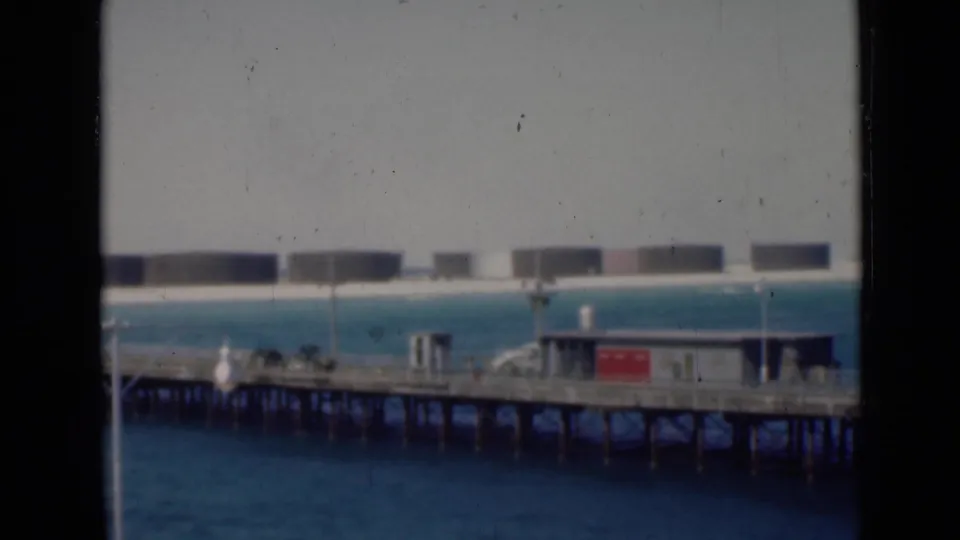 Archival 8mm view of a pier with small structures and a red sign-like panel, with large storage tanks lining the distant shore.