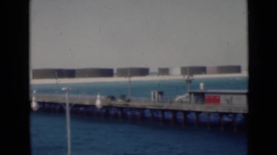 Soft-focused home-movie frame showing a wooden pier across blue water, with small structures and distant cylindrical storage tanks under a pale sky.