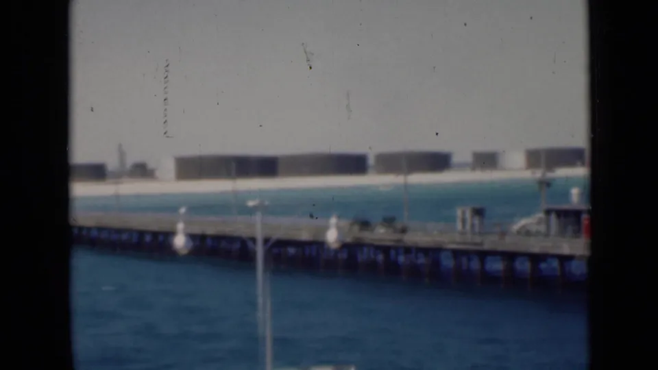 Final frame of the 1947 clip, showing a waterfront pier, light posts, small buildings, and a distant row of storage tanks across blue water.