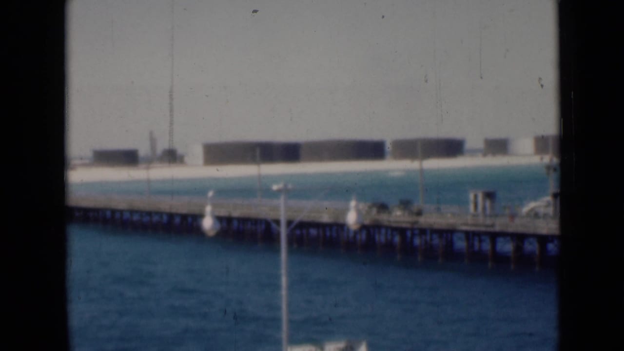 Long wooden pier over blue water with pale shoreline and distant tank-like structures.