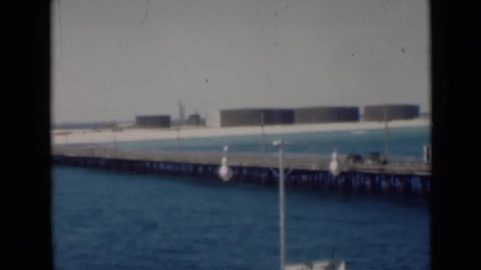 Vessel-shot 8mm frame of a waterfront pier and pale shore, partially divided by dark window-frame edges.