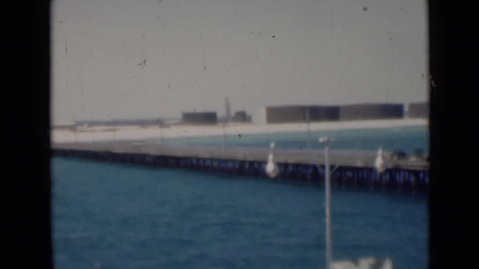 Soft 8mm frame of a pier extending across blue water toward a pale shoreline with distant industrial-looking forms.