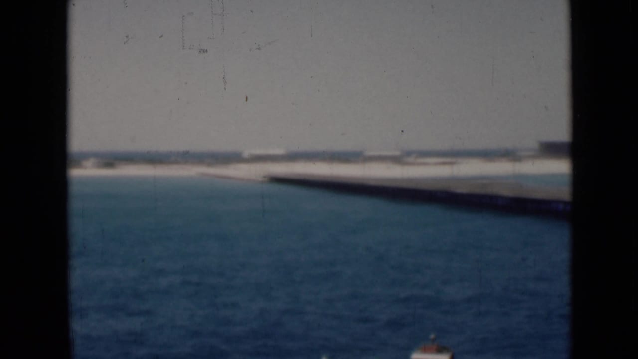 Boat-deck view over blue water toward a pale shoreline and long pier.
