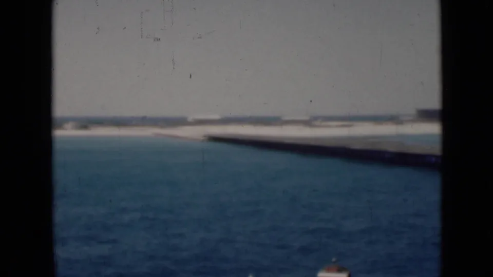 1940s 8mm boat-deck view across blue Persian Gulf water toward a pale shoreline and long pier-like structure.