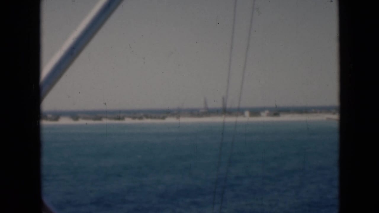 Boat-deck point of view across Persian Gulf water, with dark gate edges and a diagonal railing framing the distant shore.