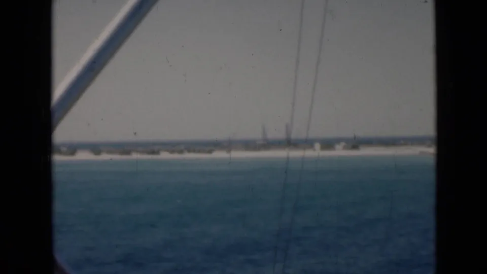 Boat-deck point of view across Persian Gulf water, with dark gate edges and a diagonal railing framing the distant shore.