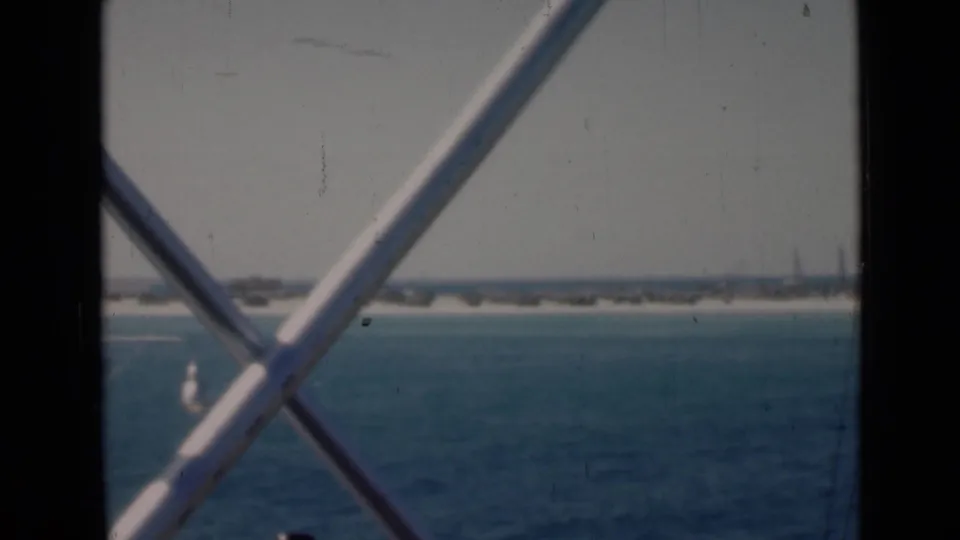 Crossed deck railings frame blue Persian Gulf water, a pale shoreline, and faint low structures in the distance.