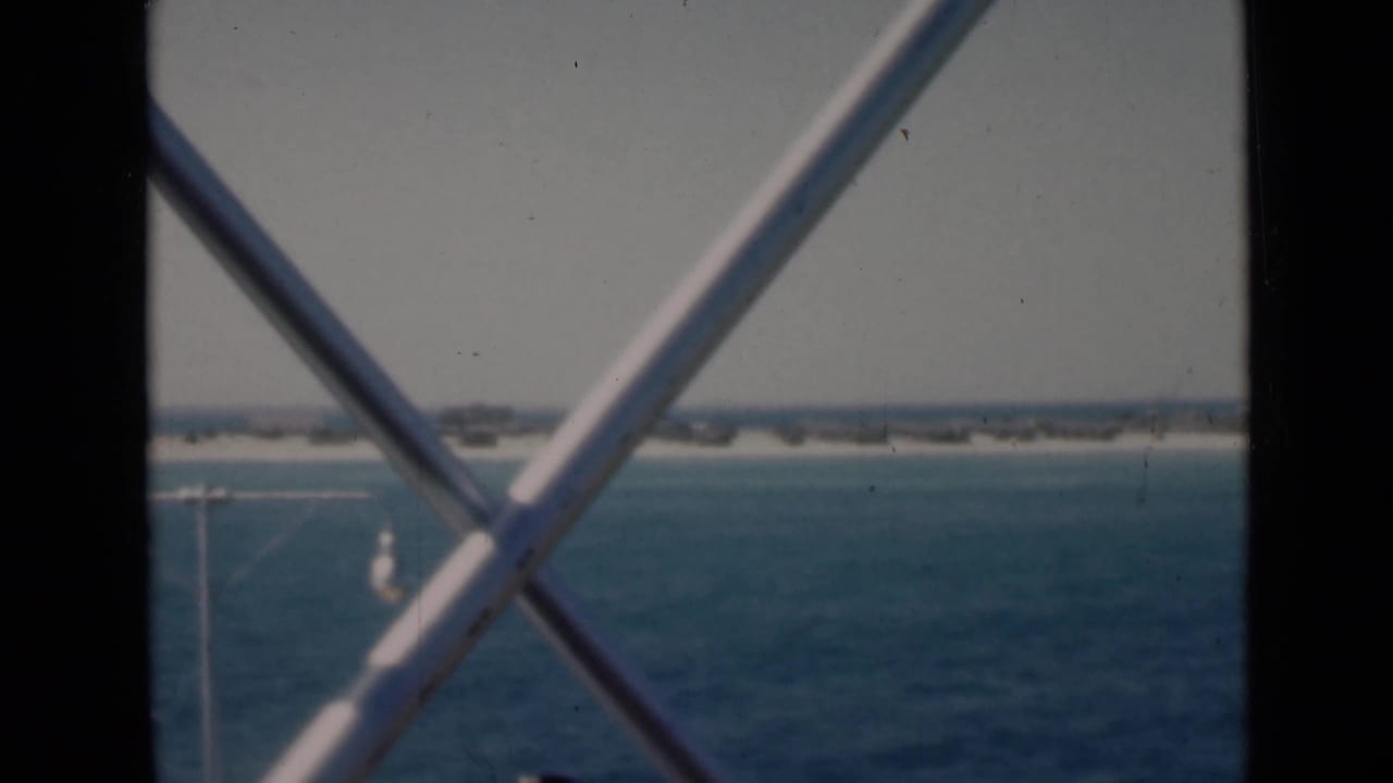 Archival 8mm view of the Persian Gulf shoreline through white steel cross-bracing, with hazy sky and open water beyond.