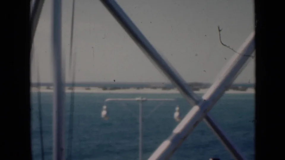 A shipboard or pier-side 8mm view looks past white metal bracing toward Persian Gulf water, a light pole, and a distant sandy shore.