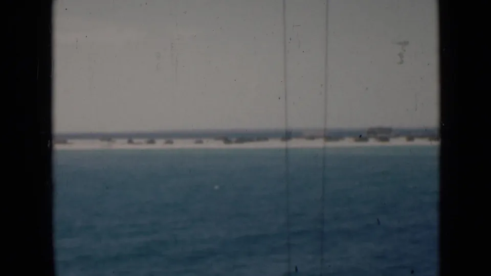 1940s 8mm frame showing blue water, a hazy distant coast, and dark vessel-window framing with visible scratches and softness.