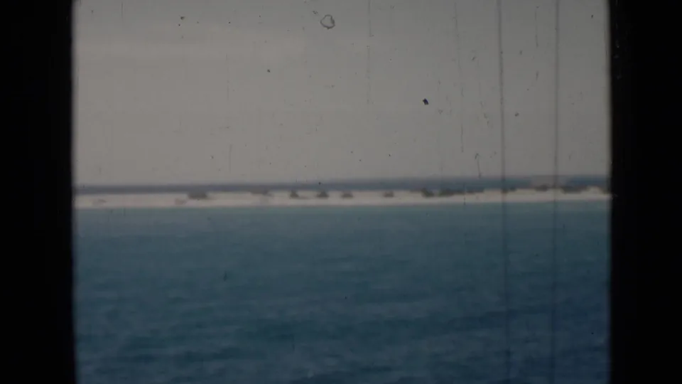 Final 8mm view of Persian Gulf water and a low shoreline under a pale sky, with dark window edges and light film scratching visible.