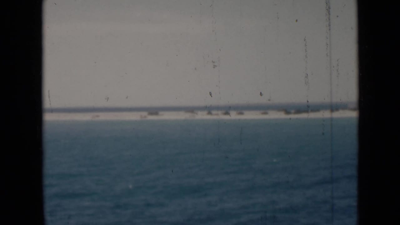 Archival 8mm view of open water under a pale sky, framed by dark window or film edges with a low distant shoreline.