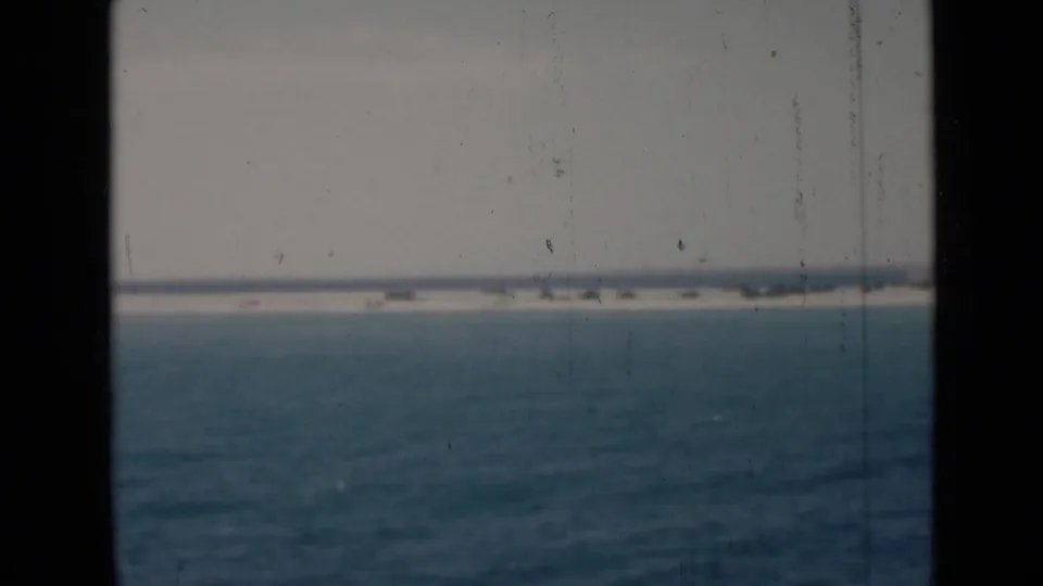 Archival 8mm view of open water under a pale sky, framed by dark window or film edges with a low distant shoreline.
