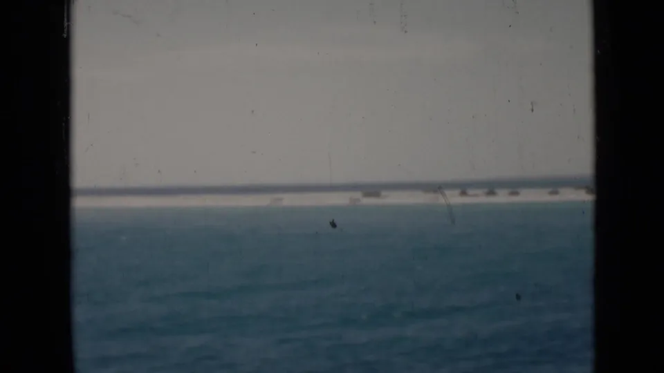 Open water and a low distant sandbar or shoreline are visible beyond a dark vertical frame strip in archival 8mm footage.