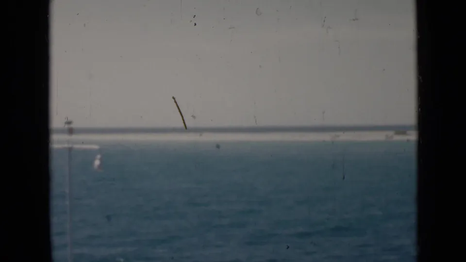 The frame holds open sea, a pale horizon, and low distant land, with dust and scratches visible in the archival 8mm scan.