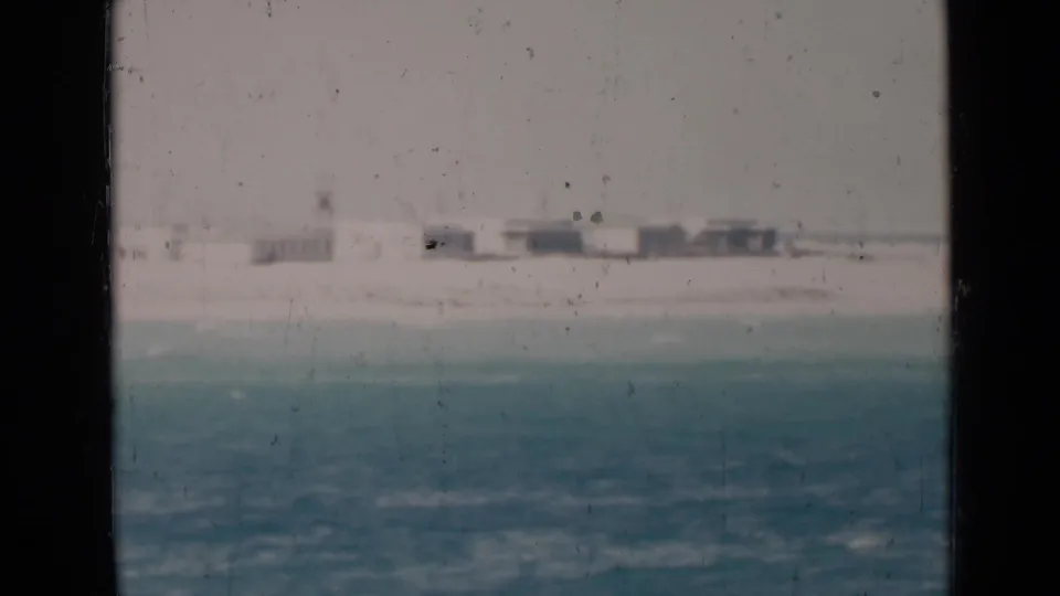 Soft 8mm view of turquoise water and a pale shoreline with low buildings in the distance, framed by dark film borders.
