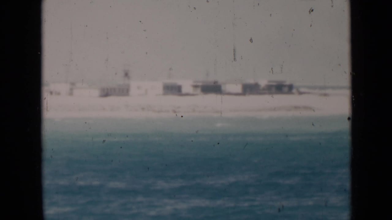 A hazy coastal view shows blue-green water, a sandy strip, and distant low-rise structures along the shore.
