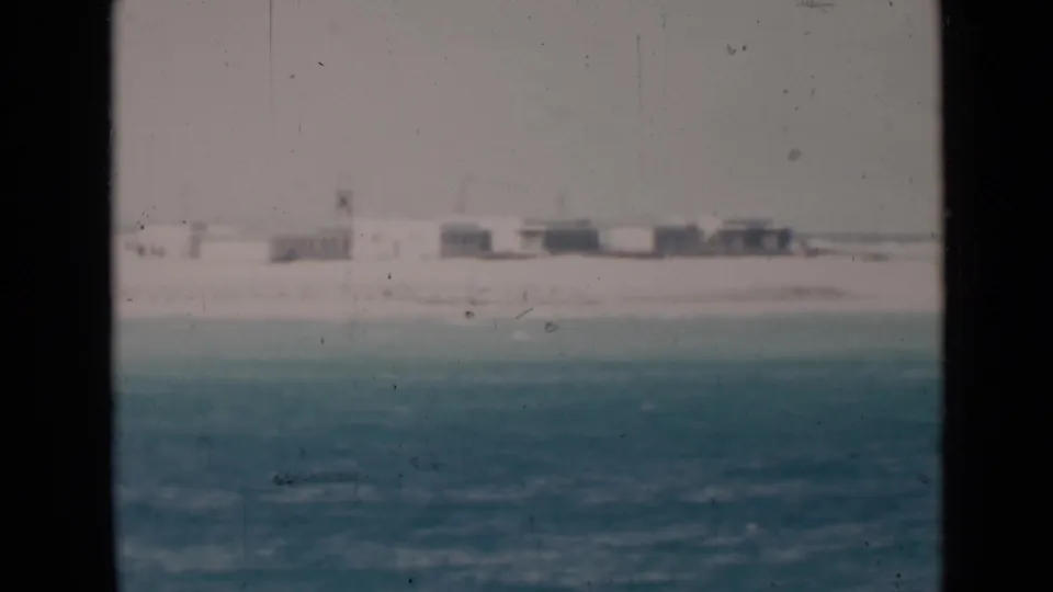 The frame shows a pale beach and small distant structures beyond textured water, with soft focus and visible film wear.