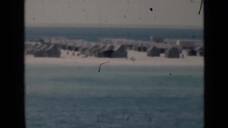 A slightly closer 8mm frame shows clustered low buildings along a sandy coast, with sea and haze beyond.