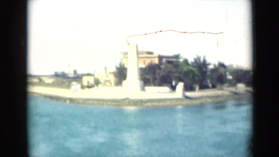 1946 Suez Canal home-movie frame showing a white waterfront monument, promenade, trees, and buildings from a moving boat.