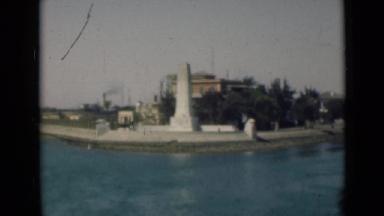 Suez Canal, Egypt frame with the obelisk-like monument centered near the water, backed by dark trees and low structures.