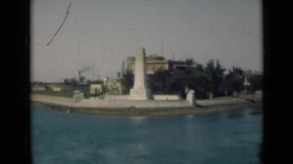 Suez Canal, Egypt frame with the obelisk-like monument centered near the water, backed by dark trees and low structures.