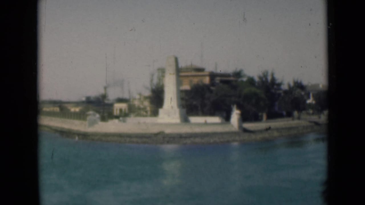 1940s Egypt canal-side view showing a white monument on the promenade with trees, buildings, and distant industrial smoke.