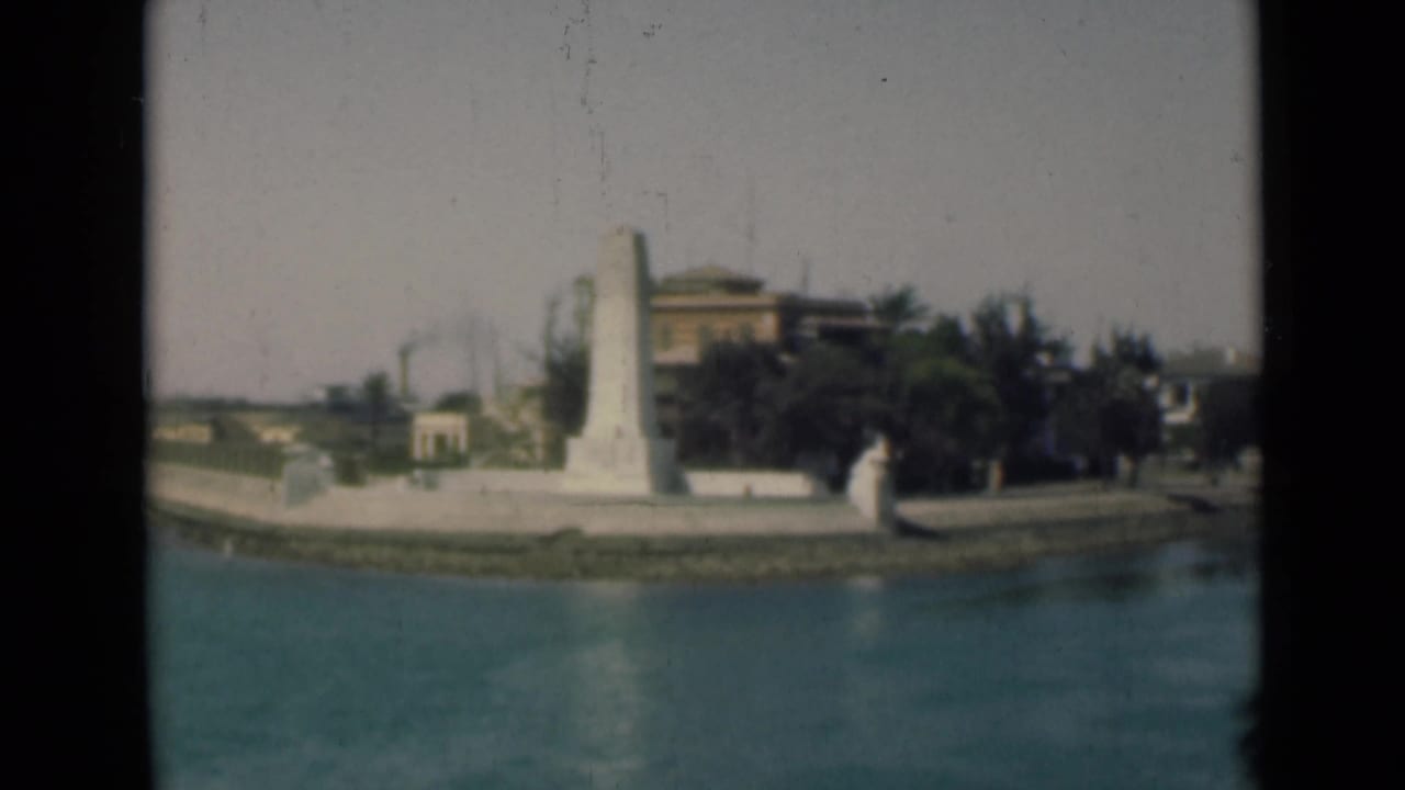 Final 1946 Suez Canal frame showing the white waterfront monument, blue water, greenery, and background buildings in soft 8mm color.