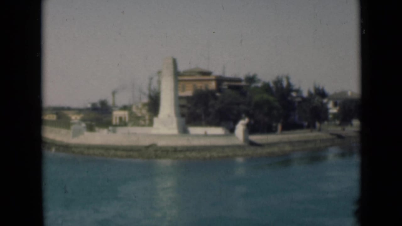 1946 8mm view along the Suez Canal showing calm water, a white monument, and low shoreline buildings in Egypt.