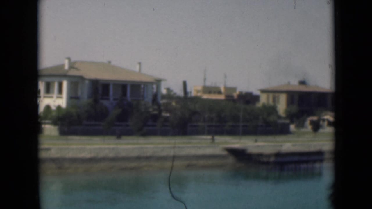1946 Suez Canal home-movie frame with low houses, garden planting, and still water in the foreground.