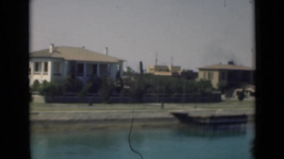 1946 Suez Canal home-movie frame with low houses, garden planting, and still water in the foreground.