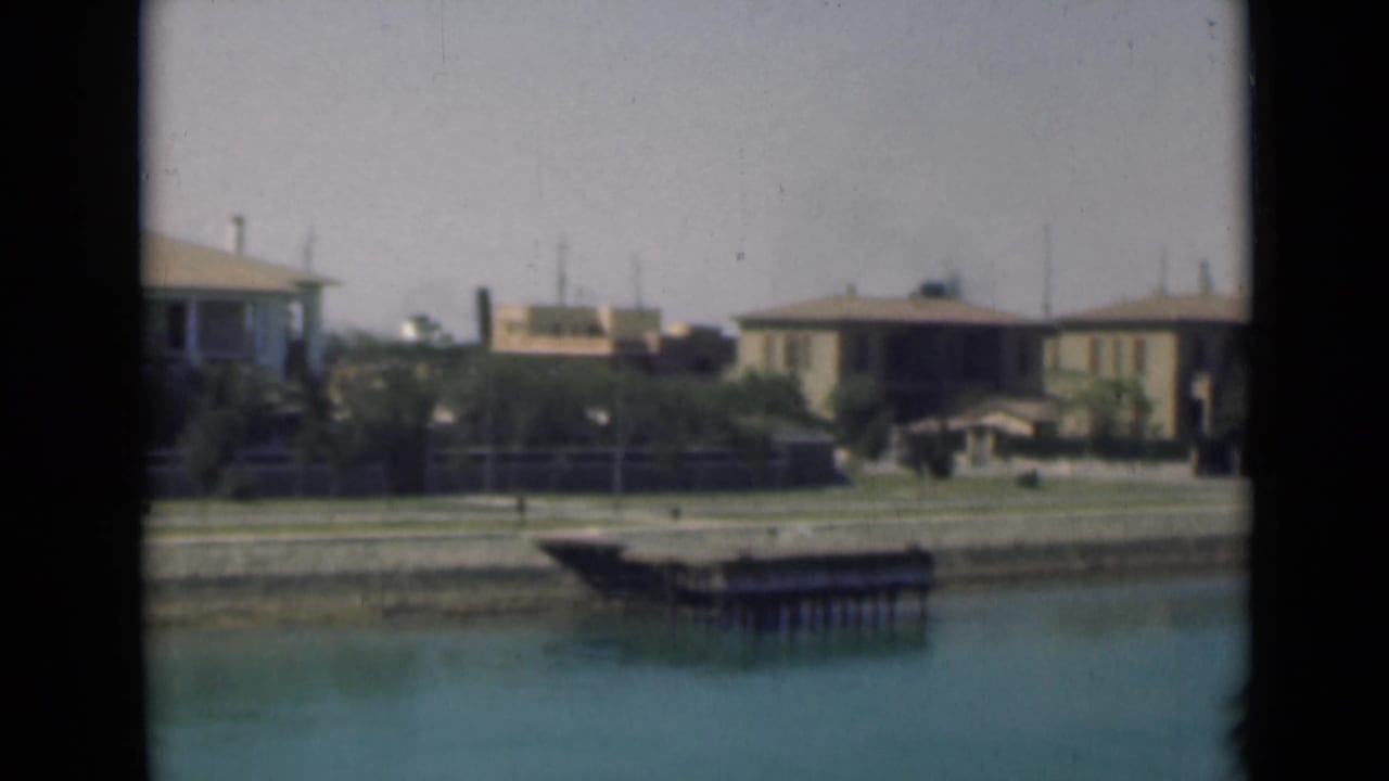 Mid-1940s 8mm Suez Canal view with a small dock, waterfront houses, shrubs, and pale sky.