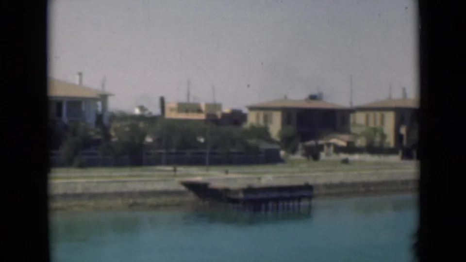 Mid-1940s 8mm Suez Canal view with a small dock, waterfront houses, shrubs, and pale sky.