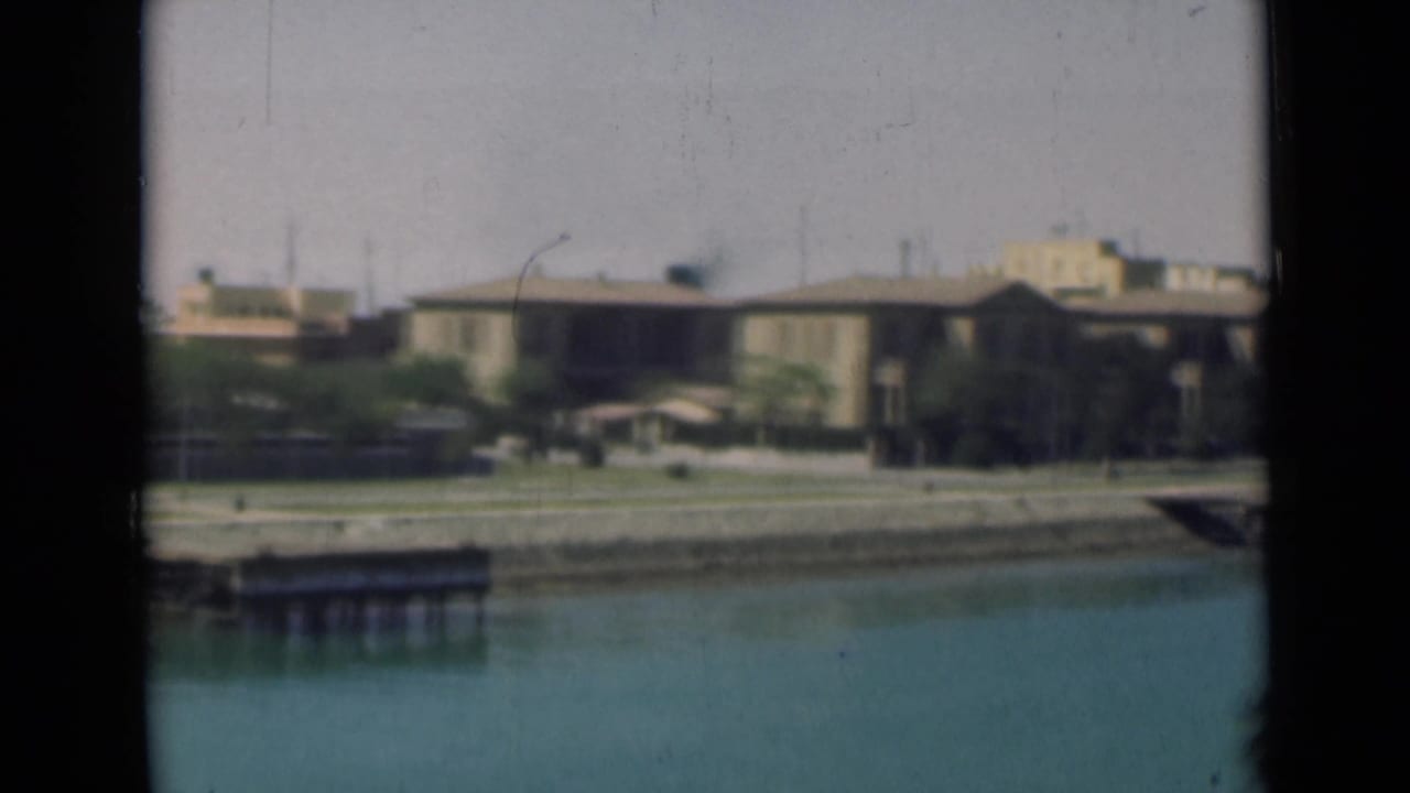 1946 Suez Canal view with calm water, a low building, palm trees, and a small dock seen through dark framing.