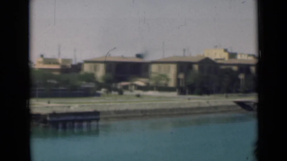1946 Suez Canal view with calm water, a low building, palm trees, and a small dock seen through dark framing.