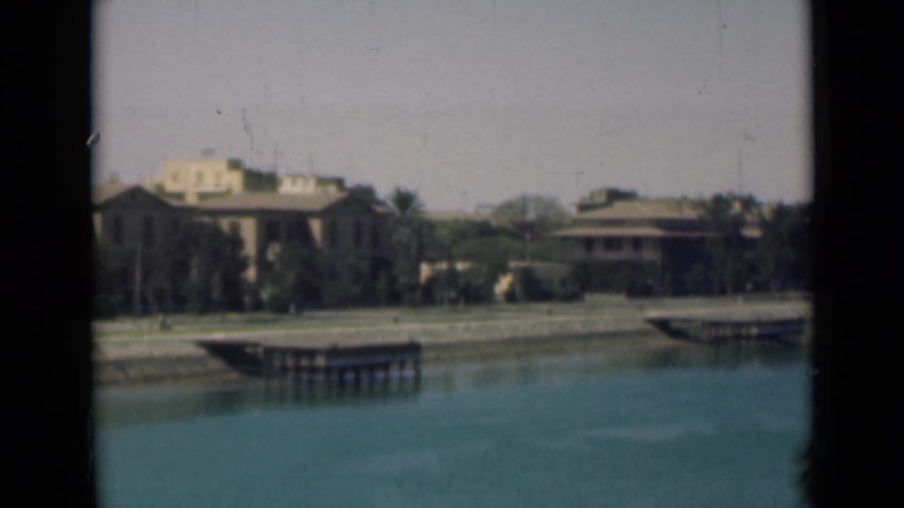 1946 Suez Canal view with calm water, low waterfront buildings, palms, docks, and pale sky.