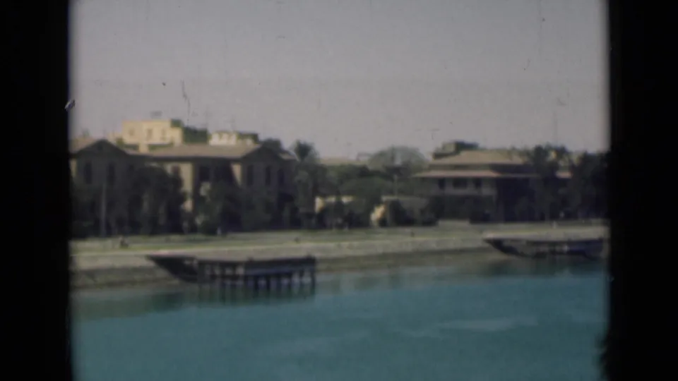 1946 Suez Canal view with calm water, low waterfront buildings, palms, docks, and pale sky.