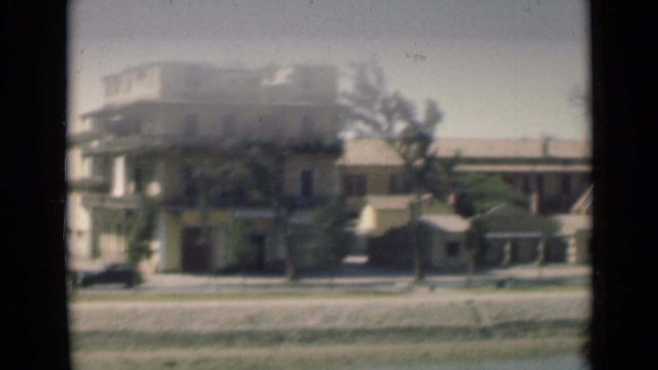 Closer 8mm view of Suez Canal-side buildings, trees, a paved embankment, and calm water, with soft focus and exposure flare.