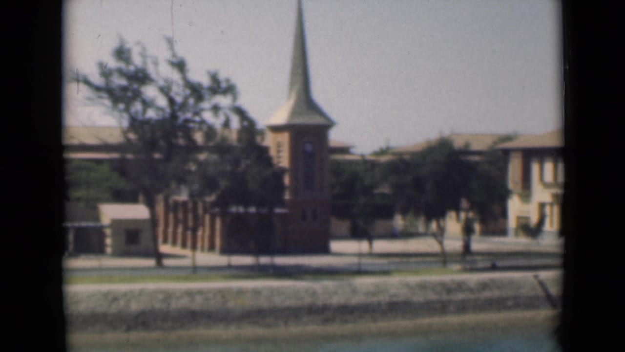 Brick church with steeple beside a pale building, trees, street, and canal-side edge.