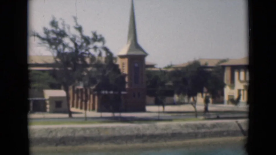 1946 Suez Canal area view of a brick church with steeple, nearby pale building, trees, and quiet street seen through dark window framing.