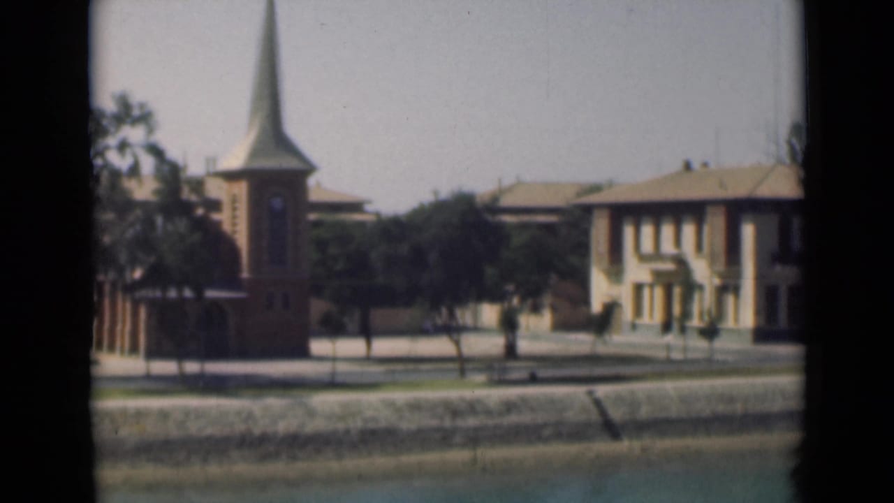 Church steeple, trees, pale building, street, and low canal-side embankment.