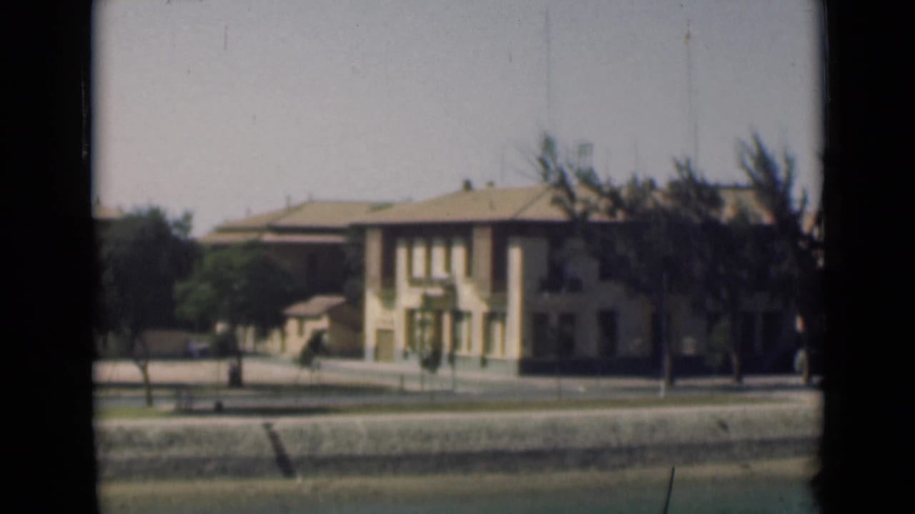 Suez Canal area exterior showing a pale multi-story building, trees, quiet street, and canal-side embankment framed by the 8mm window view.