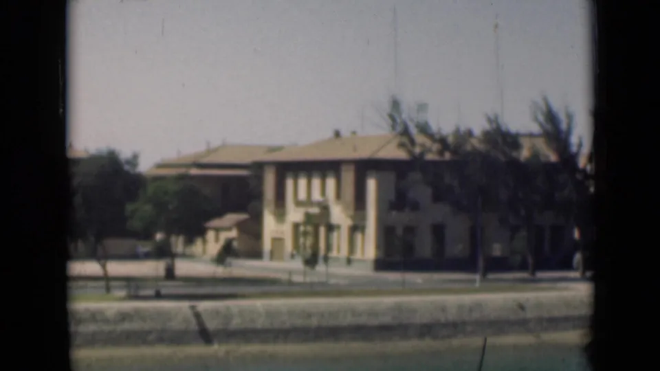 Suez Canal area exterior showing a pale multi-story building, trees, quiet street, and canal-side embankment framed by the 8mm window view.