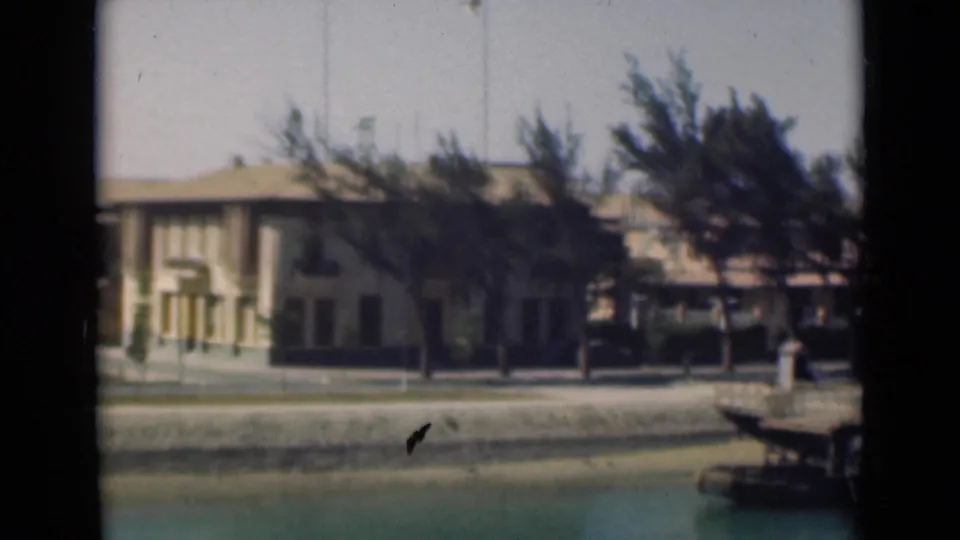 Suez Canal shoreline with a long pale building, trees, quay wall, turquoise water, and a small dock edge entering from the right.