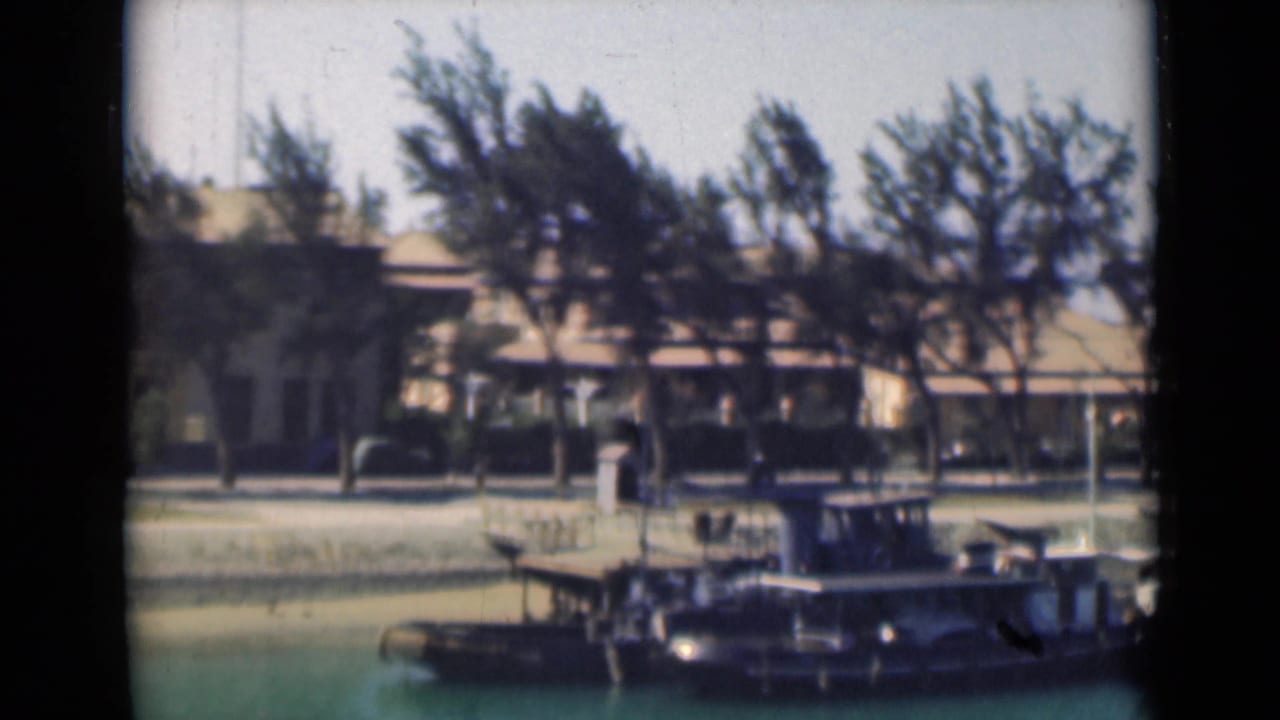 Suez Canal view with a small dark boat or docked craft in the foreground, trees along the quay, and a pale waterfront building behind.