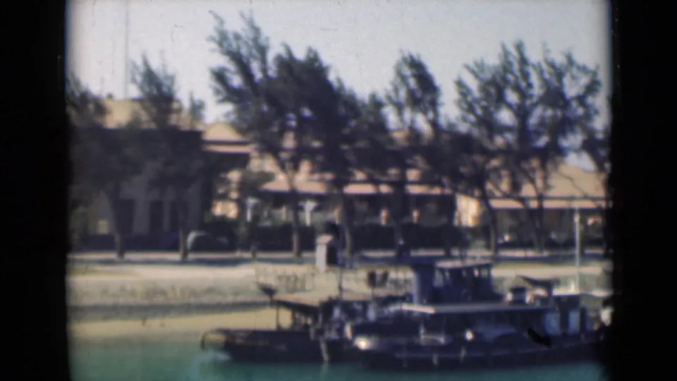 Suez Canal view with a small dark boat or docked craft in the foreground, trees along the quay, and a pale waterfront building behind.