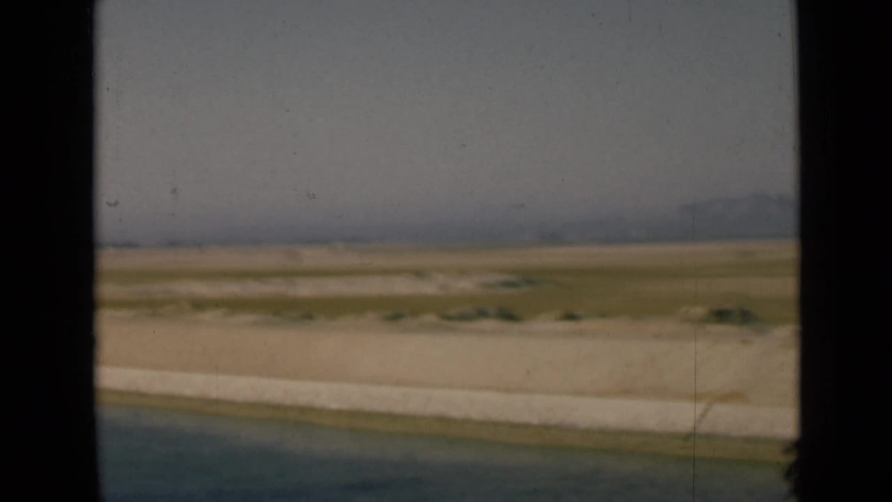 1946 Suez Canal frame showing open water, a flat canal bank, distant hazy land and pale sky from a moving home-movie viewpoint.
