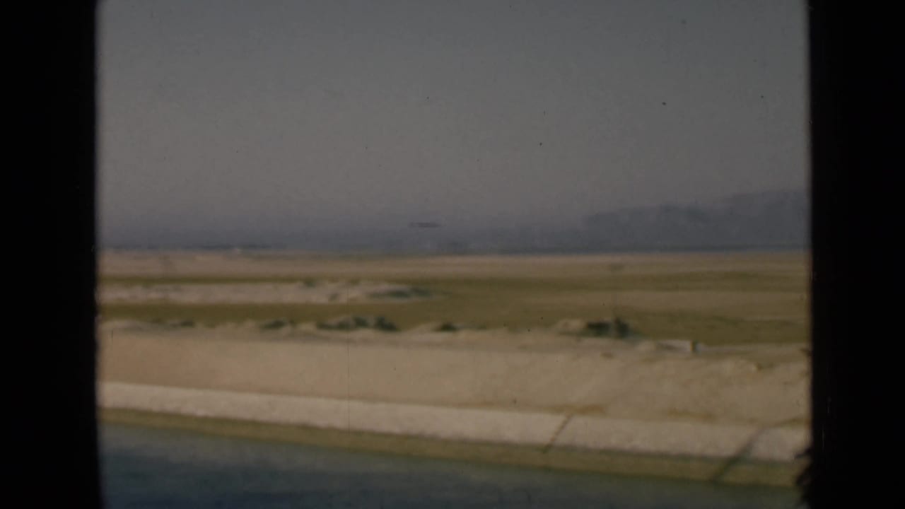 Canal edge, concrete bank, desert flats, distant hills, and hazy sky seen through dark film borders.