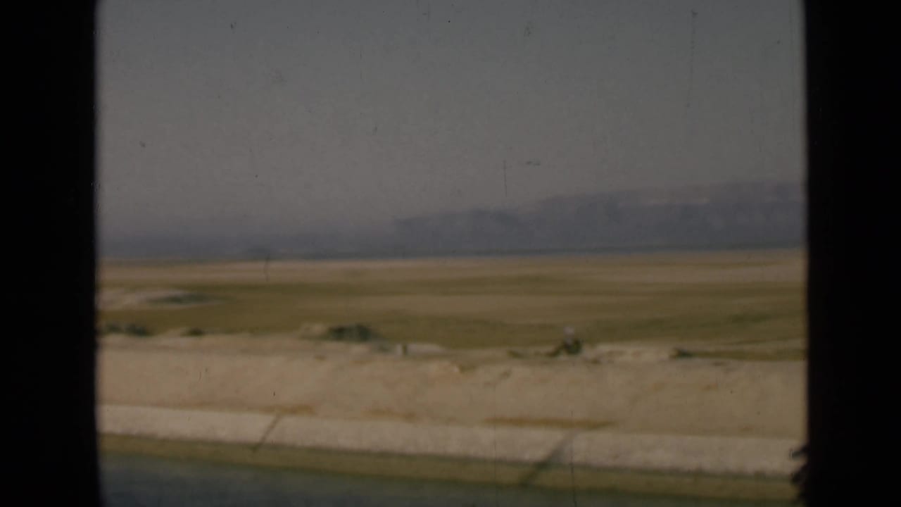 Open desert plain beside a pale embankment, with low mountains under a gray sky.